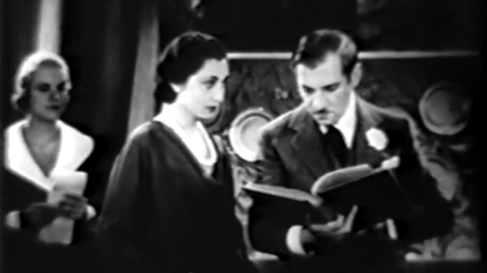 Schwarz-Weiß-Szene mit Aline MacMahon als Sprechcoach beim skeptischen Blick auf Gregory Ratoff als in ein Drehbuch vertiefter Studioboss, im Hintergrund eine wartende Mitarbeiterin.