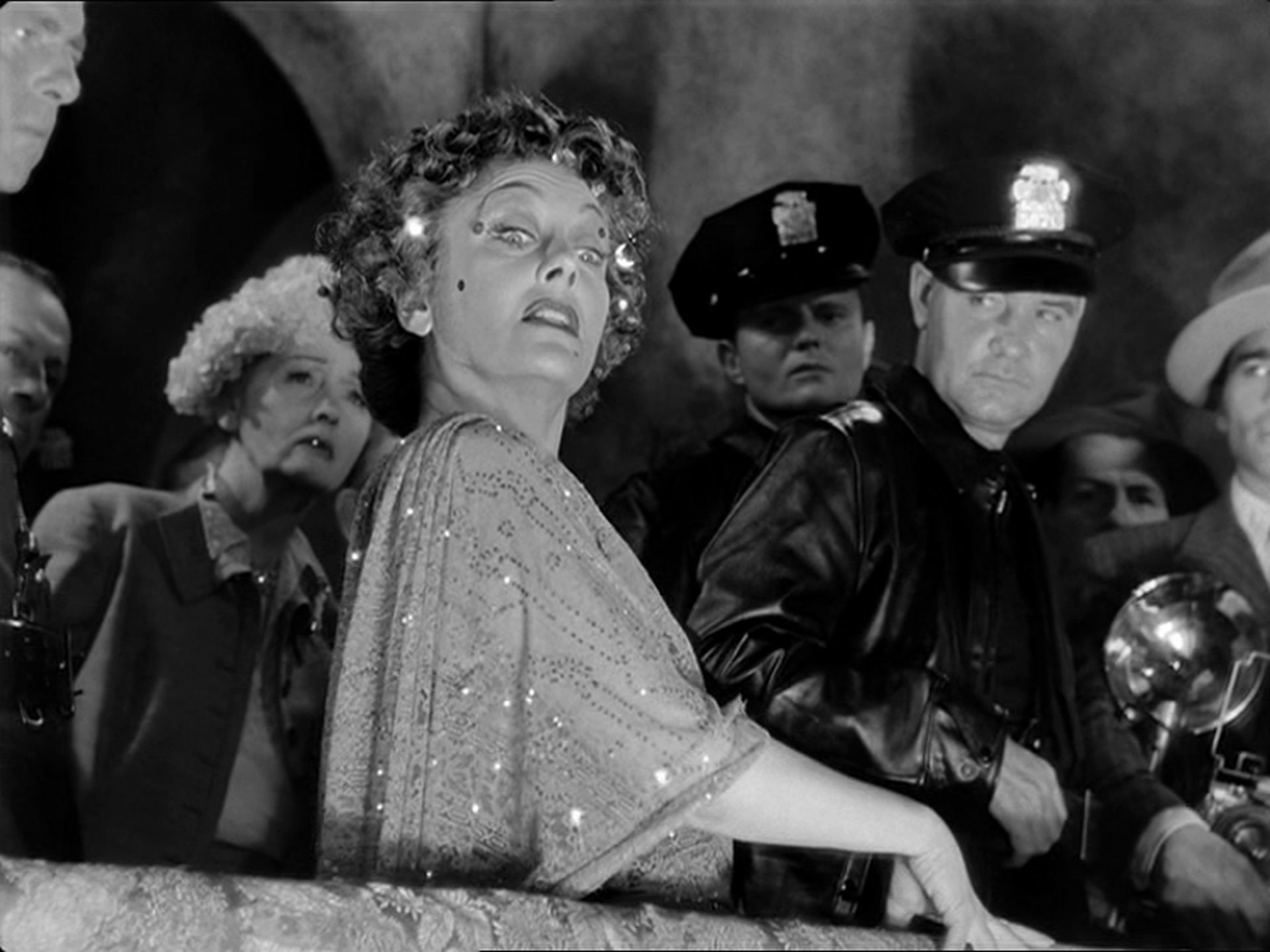 Gloria Swanson als Norma Desmond mit irrem Blick in ihrer Villa, beagrwöhnt von Polizisten im Hintergrund.