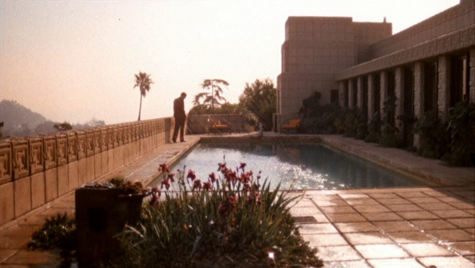 Im Hintergrund des Bildes steht ein Mann mit leicht gesenktem Kopf am Rand eines großen Pools; das am rechten Bildrand erkennbare Gebäude ist in Wirklichkeit das berühmte Ennis House in Los Angeles.