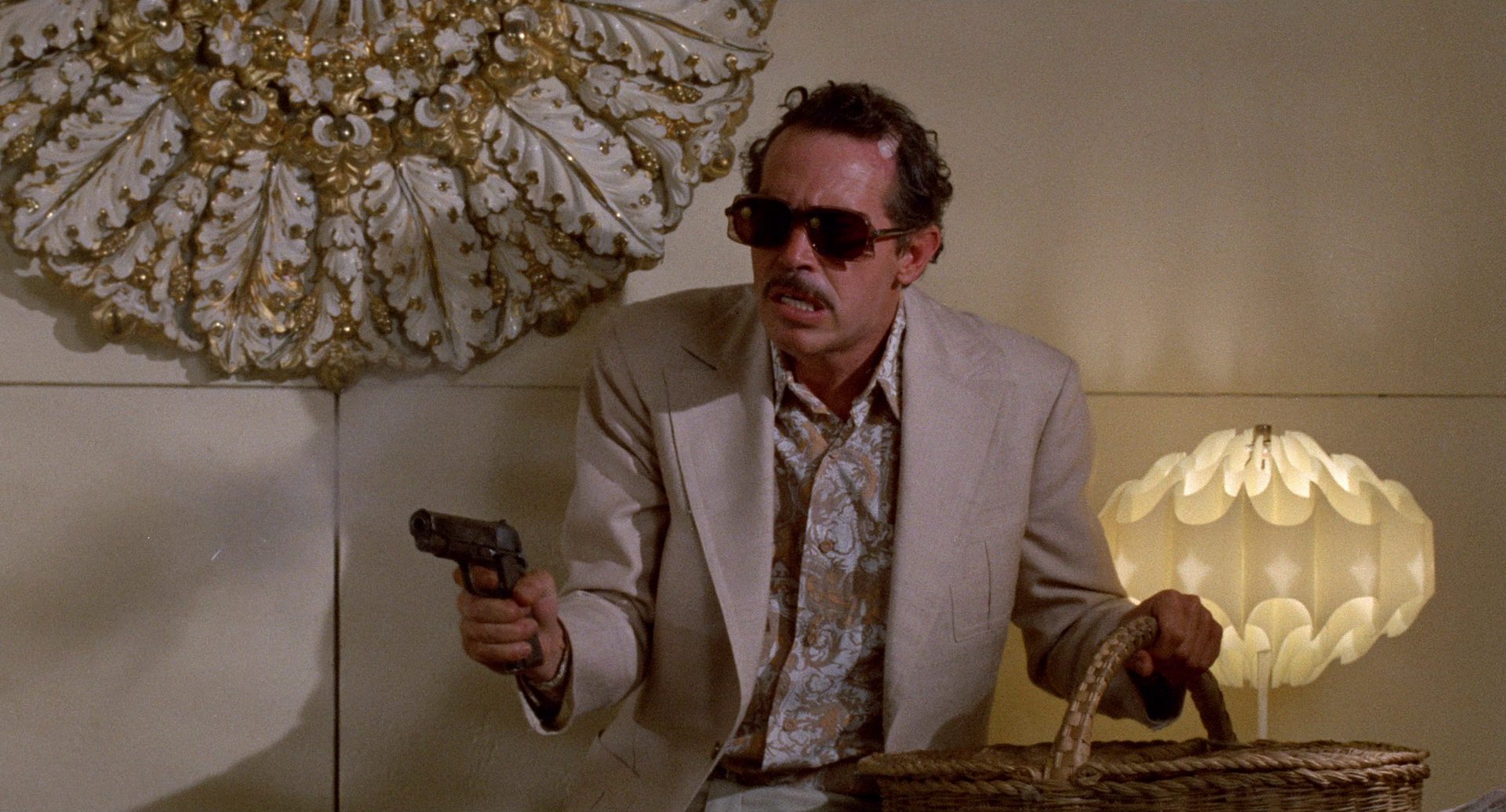 Warren Oates im hellen Sakko, mit Sonnenbrille und gezückter Pistole im Büro der Kopfgeldjäger.