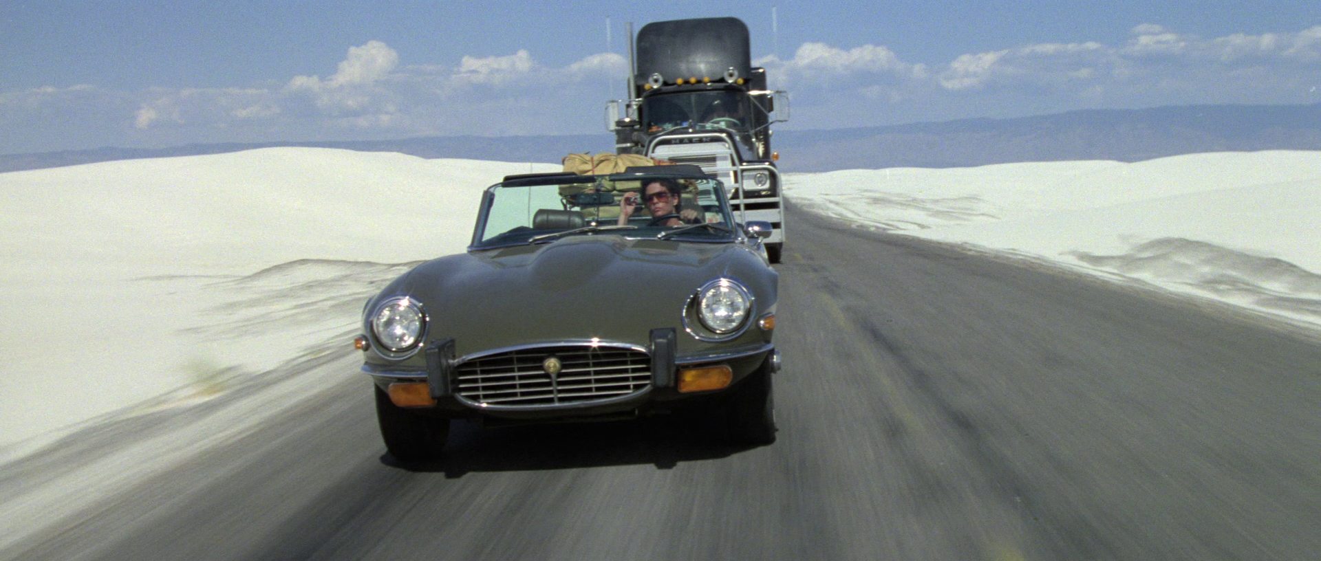 Ali MacGraw am Steuer eines Jaguar „E-Type“ auf einem Highway, hinter ihr ein Truck.