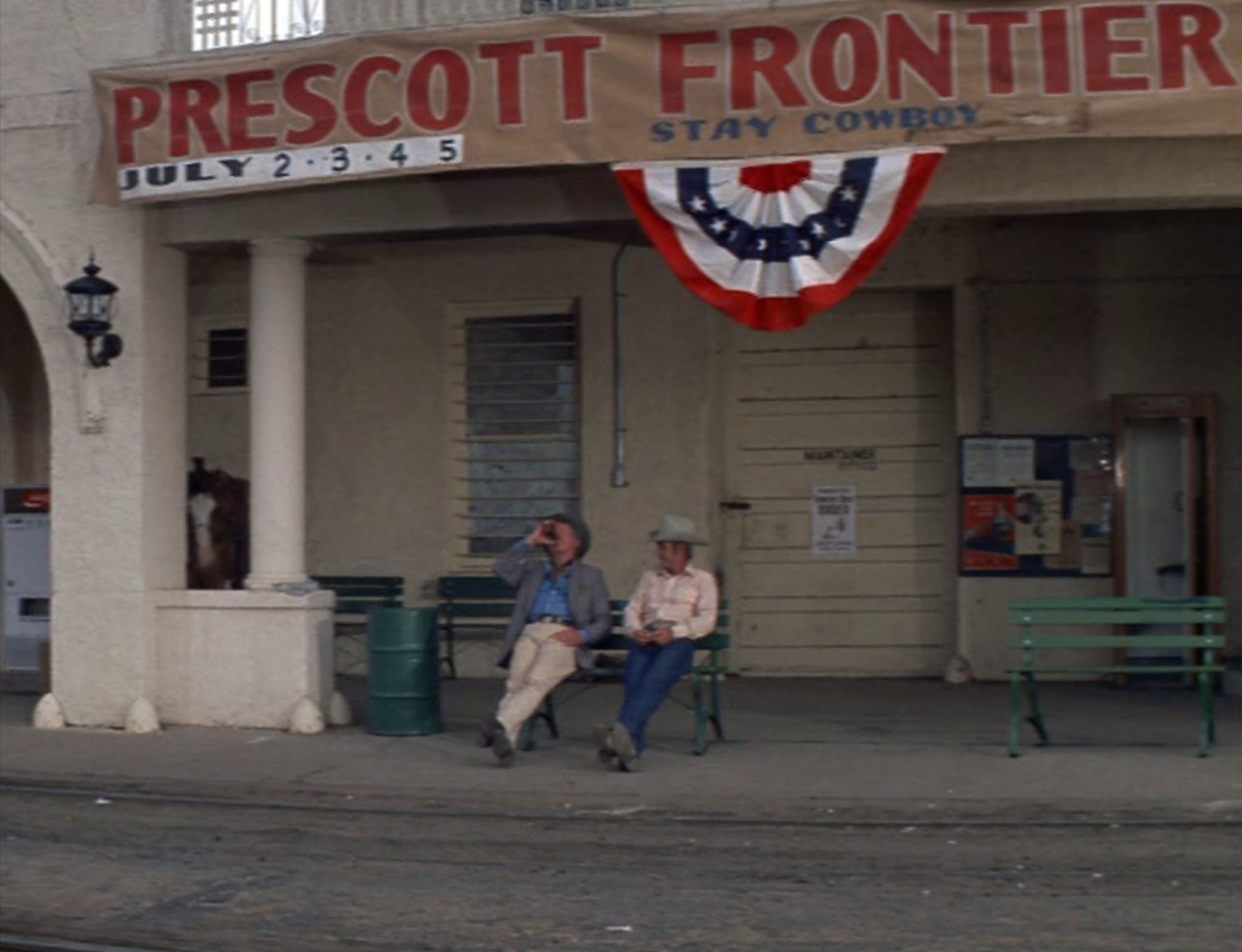 Ace und Junior Bonner aus der Distanz betrachtet auf einer Bank am Bahnhof. Über ihnen ein Banner für das „Prescott Frontier“-Festival.