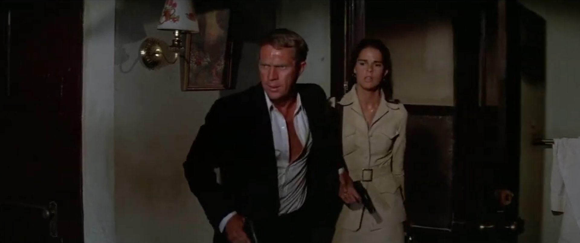 Steve McQueen und Ali MacGrac mit Pistolen bewaffnet in eilig-alerter Haltung in einem schäbigen Hotelzimmer.