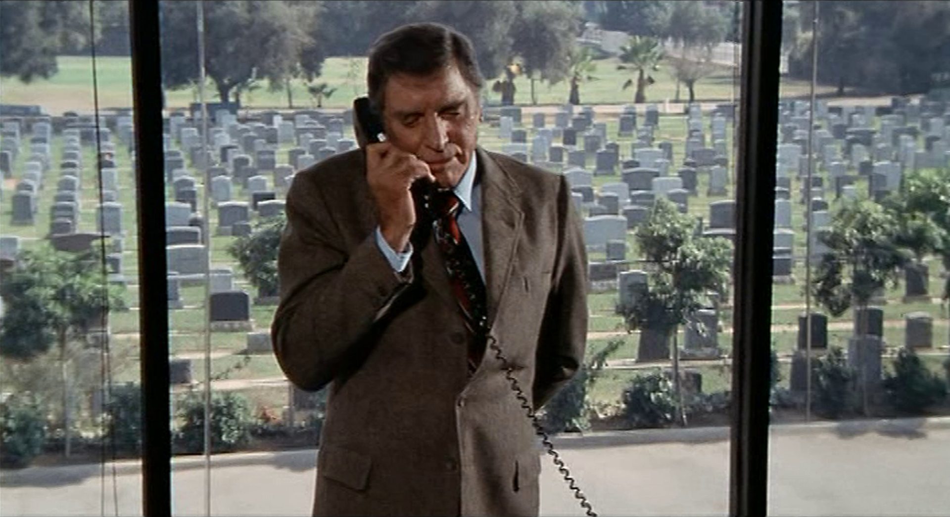 Burt Lancaster steht mit dem Telefonhörer am Ohr vor einem großen Fenster, das im Hintergrund einen Friedhof mit unzähligen Grabsteinen zeigt.