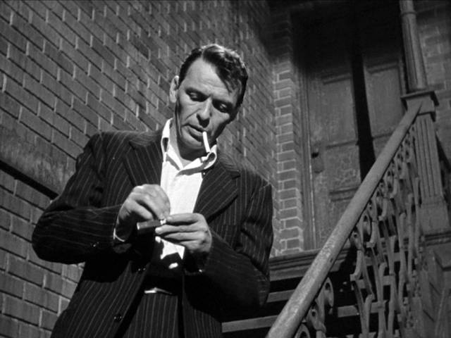 Frank Sinatra als Frankie Machine, der sich an einer Treppe eine Zigarette anzündet